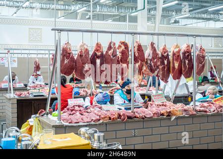 Carne cruda mostra il mercato di Altyn Orda, il più grande mercato di Almaty, Kazakhstan. Tagliare pezzi di carne appesi; venditori dietro i banconi. Foto Stock