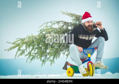 Eccitata ciclista a santa Hat con pino albero di Natale in bicicletta per bambini. Hipster felice che trasporta l'albero di natale sulla neve bianca. Uomo a cavallo triciclo su neve Foto Stock