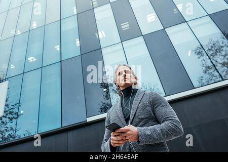 Uomo d'affari moderno con smartphone nelle sue mani su una strada della città. Bell'uomo che indossa le cuffie e parla sul telefono cellulare, ascoltando musica o podc Foto Stock