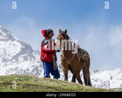 Kazbegi, Georgia - 02 maggio 2017: Un cavallo non identificato si prende cura del suo cavallo sadled sullo sfondo di vette innevate Foto Stock