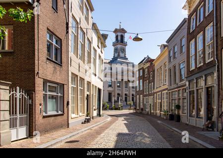 Dordrecht, Paesi Bassi-Luglio 2021; Vista su strada lastricata di Grotekerksebuurt, fiancheggiata da edificio storico e municipio con l'iconico orologio t Foto Stock