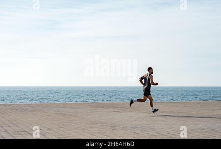 Fit giovane che corre lungo il lungomare. Corridore maschio che fa jogging la mattina presto. Atleta che pratica allenamento fitness all'aperto. Allenamento durante il blocco outsi Foto Stock