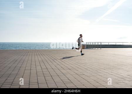 Fit giovane che corre lungo il lungomare. Corridore maschio che fa jogging la mattina presto. Atleta che pratica allenamento fitness all'aperto. Allenamento durante il blocco outsi Foto Stock