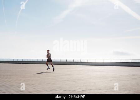 Fit giovane che corre lungo il lungomare. Corridore maschio che fa jogging la mattina presto. Atleta che pratica allenamento fitness all'aperto. Allenamento durante il blocco outsi Foto Stock