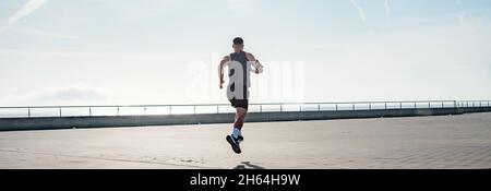 Fit giovane che corre lungo il lungomare. Corridore maschio che fa jogging la mattina presto. Atleta che pratica allenamento fitness all'aperto. Allenamento durante il blocco outsi Foto Stock