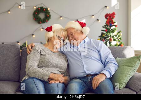 Sorridente coppia anziana amorevole in Natale cappellini festive seduti sul divano e abbracciato durante Natale Foto Stock