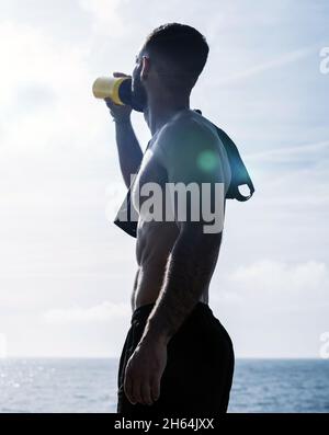 Giovane uomo in forma che beve dell'acqua da una bottiglia e che riposa dopo l'addestramento all'aperto. Allenamento mattutino Foto Stock