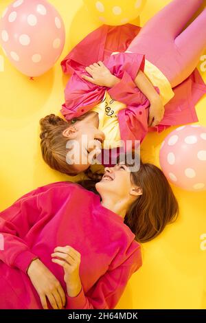2 ragazze sorelle che si trovano su sfondo giallo in studio guardando l'un l'altro e sorridendo tra palloncini felice concetto di infanzia, rapporto tra Foto Stock
