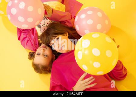 2 ragazze sorelle sono sdraiati su sfondo giallo in studio guardando la macchina fotografica tra palloncini, concetto di infanzia, rapporto tra bambini in famiglia Foto Stock