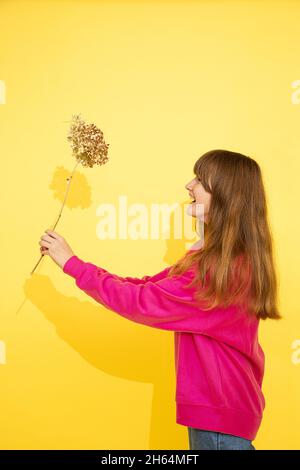 foto luminosa di ragazza adolescente con capelli lunghi e scuri in maglione rosa con fiore in mano su sfondo giallo. tiene il fiore nelle mani, ridendo felicemente a. Foto Stock