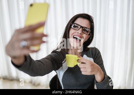 Ritratto di una ragazza latina allegra vestita con vestiti grigi che tiene una tazza di caffè gialla mentre prende un selfie a casa Foto Stock