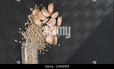 Aglio e pepe. Garlics naturale e pepe bianco in bottiglia di vetro su fondo nero di tappetino. Ingrediente per molte ricette alimentari del mondo. Repressone Foto Stock