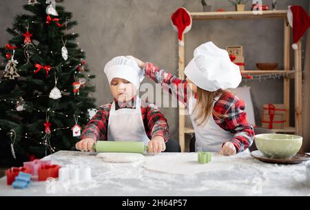 i bambini preparano i biscotti di natale, il ragazzo arrotola l'impasto, la sorella lo aiuta raddrizzando il cappello, cucinando il pan di zenzero insieme, sfondo festoso della parete. Foto Stock