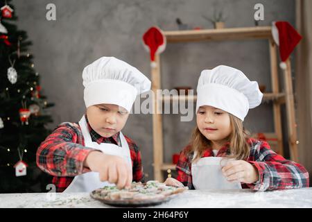 I bambini preparano biscotti di Natale e pan di zenzero. Il ragazzo decora i biscotti con glassa colorata. Fratello e sorella nella divisa del Baker Foto Stock