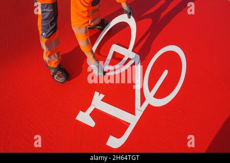 10 novembre 2021, Hessen, Francoforte sul meno: Un operaio inserisce un simbolo di bicicletta riflettente nella vernice rossa fresca. Una pista ciclabile è contrassegnata con vernice rossa a Gartenstraße/Walter-Kolb-Straße. Foto: Andreas Arnold/dpa Foto Stock