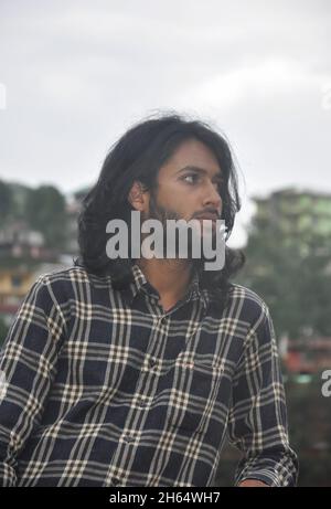 Vista frontale di un bell'uomo dall'aspetto sud asiatico dai capelli lunghi e dal bearded giovane che guarda lateralmente mentre si alza all'esterno Foto Stock