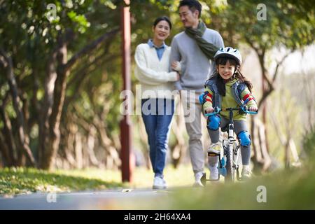 bambina asiatica con casco e completo di protezione in bicicletta nel parco della città con i genitori che guardano da dietro Foto Stock