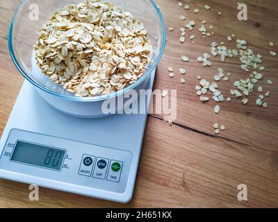 OAT groats, grits pesati in vaso di vetro su bilance elettroniche da cucina su tavola di legno. Foto Stock
