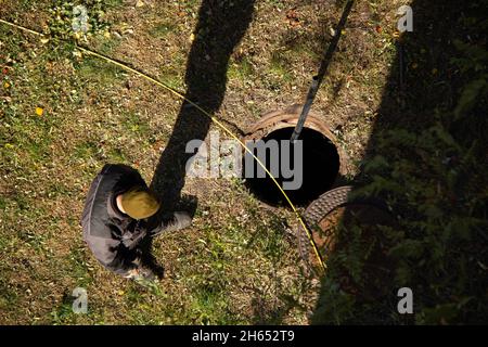 Operaio in un uniformato vicino ad una botola di fognatura aperta. Cavo elettrico giallo e tubo sullo sfondo del prato autunnale. Lavori di ristrutturazione di un impianto idraulico Foto Stock