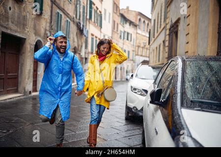 Una giovane coppia allegra di impermeabili corre dalla pioggia verso l'auto, mentre ha fatto una passeggiata in città in un'atmosfera piovosa. Passeggiata, pioggia, città, rela Foto Stock