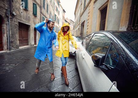 Una giovane coppia innamorata sta per andare in macchina mentre si fa una passeggiata in città in impermeabile in un'atmosfera allegra durante una giornata piovosa. Cammina, pioggia, cit Foto Stock