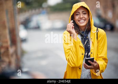 Ragazza sorridente in un impermeabile giallo ascolta la musica sotto la pioggia Foto Stock