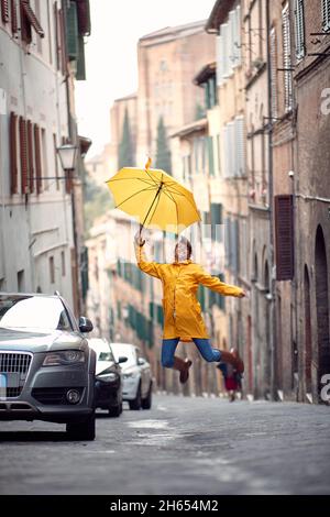 Una giovane ragazza con un impermeabile giallo e un ombrello salta sulla strada mentre si gode una passeggiata attraverso la città in modo allegro in una giornata piovosa. Foto Stock