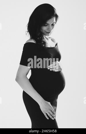 Bella giovane donna incinta che abbraccia il ventre in posa in studio, foto in bianco e nero. Buona gravidanza e maternità Foto Stock