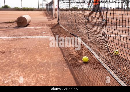 Palle da tennis vicino alla rete con rullo pesante e persona di manutenzione su campo di argilla in giornata di sole all'aperto. Concetto di allenamento sportivo Foto Stock