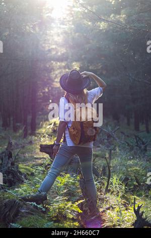 Donna escursionistica con cappello e zaino che guarda al sole nella foresta. Il viaggiatore femminile è in piedi in fascio di sole a Woodland. Divertimento della natura Foto Stock