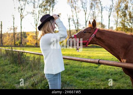 Cowgirl che gioca con il suo giovane cavallo in fattoria. Friedship tra persone e animali domestici. Curioso fallo e donna che indossa cappello cowboy e maglia swe Foto Stock