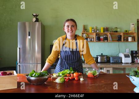 Ritratto di donna sorridente indossando grembiule appoggiato sul bancone della cucina con varie verdure fresche Foto Stock