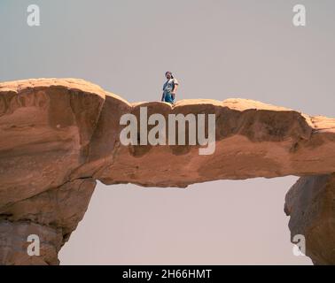 Uomo turista in piedi su Um Frouth Rock Bridge, un alto ponte di roccia naturale nel deserto di Wadi Rum, Giordania. Foto Stock