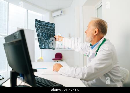 Vista laterale di un medico adulto maschio esperto che analizza l'immagine della colonna vertebrale MRI del paziente seduto alla scrivania con il computer nella stanza dell'ufficio medico. Foto Stock