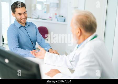 Vista dal retro a paziente giovane allegro ascolto medico maschile senior dando assistenza sanitaria in ospedale. Foto Stock