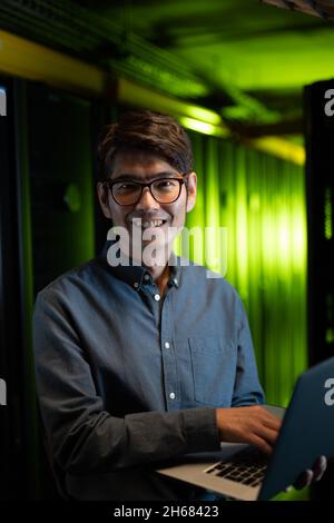 Ritratto di ingegnere asiatico maschile sorridendo mentre si usa il computer portatile in sala server Foto Stock