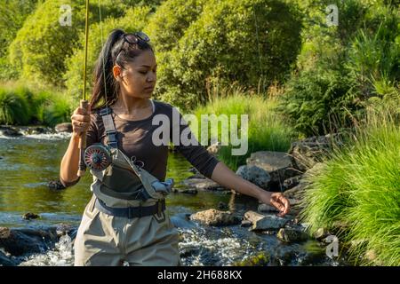 Una giovane donna asiatica in piedi pesca a mosca in un riffle su un fiume Foto Stock