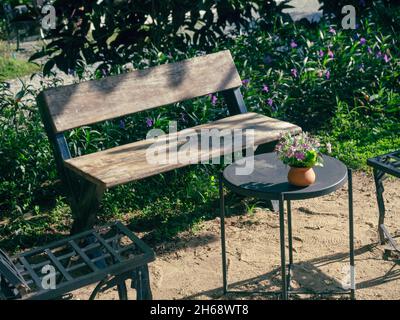 Pentola in terracotta con decraotion di fiori su tavola rotonda nera con panca in legno antico e di color empty in giardino al mattino rilassato con sole. Foto Stock