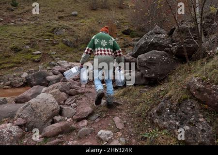 Un alpinista, uomo adulto anziano, di circa 70 anni, indossando un maglione di lana, cammina lungo un percorso tra le rocce, verso un flusso d'acqua, trasportando Foto Stock