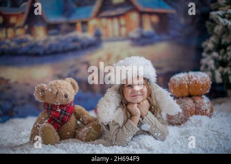 Una ragazza in un caldo cappello bianco d'inverno si trova sul suo stomaco vicino ad un cucciolo di orso giocattolo sullo sfondo di un villaggio d'inverno Foto Stock