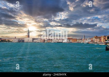 Acqua di mare superficie con paesaggio urbano e sole che brilla attraverso cielo nuvoloso Foto Stock