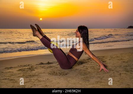 Bella donna che fa pilates esercizio con palla sulla spiaggia al tramonto. Donna seduta sulla sabbia facendo pilates esercizio tenendo palla morbida tra sollevato Foto Stock