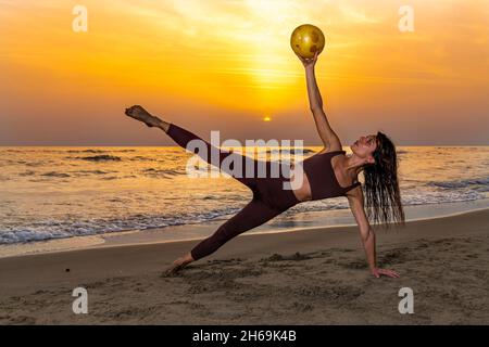 Bella donna che fa pilates esercizio con palla sulla spiaggia al tramonto. Donna che tiene una palla morbida con il braccio sollevato mentre lei solleva la gamba. Concep Foto Stock