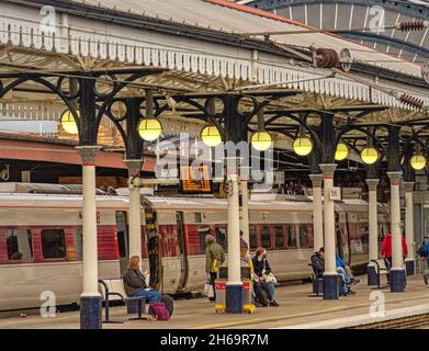 Piattaforma della stazione ferroviaria con i passeggeri in attesa di un treno. In alto si trova una tettoia storica con colonne. I carrelli sono sullo sfondo. Foto Stock