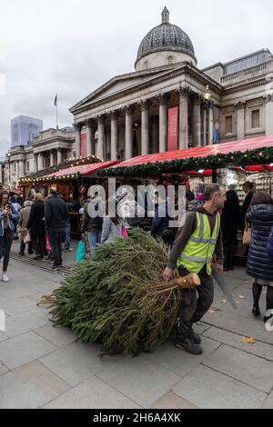 Un lavoratore porta un albero di Natale al mercato di Natale, Trafalgar Square, all'inizio della stagione festiva 2021 Natale nella capitale, Londra, Inghilterra. Foto Stock