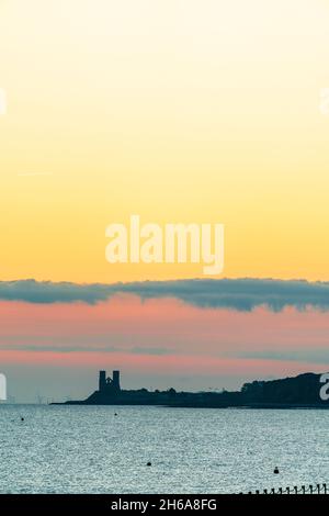 Cielo rosso e giallo dell'alba con alcune nuvole di cumulo mediocri sopra la lontana rovina delle torri gemelle della chiesa anglosassone a Reculver sulla costa del Kent. Foto Stock