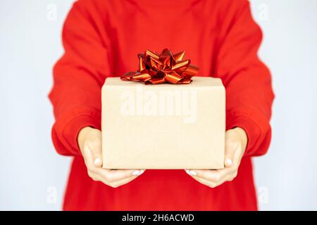 Regali nelle mani della donna. Consegna espressa. Una giovane donna in una felpa rossa tiene una scatola di cartone con un arco rosso. Shopping online sale. Quarantena Foto Stock