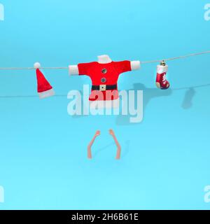 Blusa Babbo Natale, cappello rosso con pompon bianco e una scarpa rossa appesa su una corda. Braccia donne sotto. Sfondo blu pastello. Natale surreale o New Ye Foto Stock
