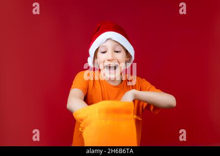 Un ragazzo in un cappello di Santa rummages in un sacchetto giallo del regalo di Natale e ride ad alta voce Foto Stock