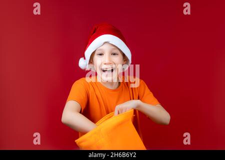 Un ragazzo in un cappello di Santa rummages in un sacchetto giallo del regalo di Natale e ride ad alta voce Foto Stock